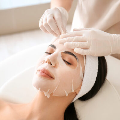 Cosmetologist applying sheet mask on woman's face in beauty salon